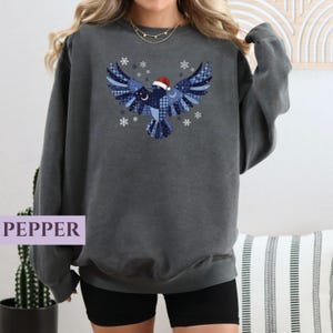 May include: A dark gray sweatshirt featuring a blue bird design with a Santa hat and snowflakes. The bird is composed of various blue plaid and patterned fabrics. The word "PEPPER" is printed in a purple box at the bottom left.