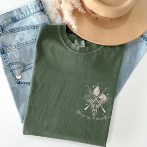 May include: An olive green t-shirt with a graphic design featuring a deer, crossed arrows, and howling wolves. The text "Always up to something" is below the graphic. A tan hat and blue jeans are also in the image.