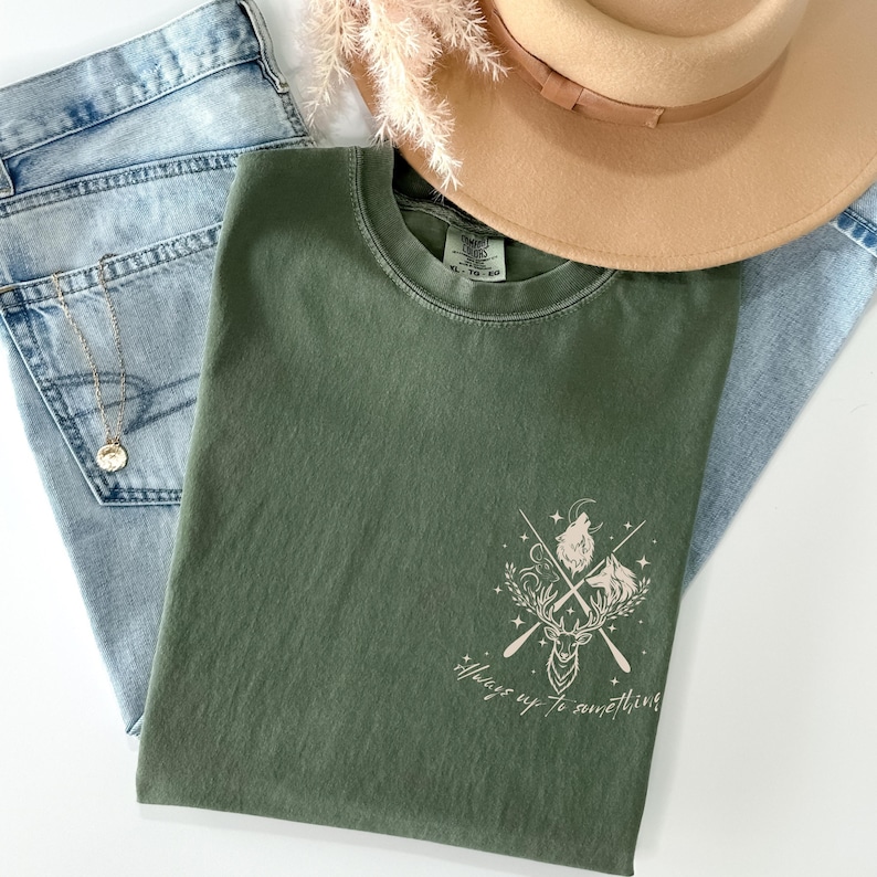 May include: An olive green t-shirt with a graphic design featuring a deer, crossed arrows, and howling wolves. The text "Always up to something" is below the graphic. A tan hat and blue jeans are also in the image.