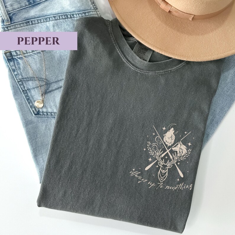 May include: A dark grey t-shirt featuring a deer, wolf, and crossed oars graphic, with the text "Always up to something." The shirt is on light blue jeans, a gold necklace, and a beige hat.