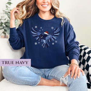 May include: A navy blue sweatshirt featuring a patchwork bird design with a Santa hat and snowflakes. The text "TRUE NAVY" is displayed on a rectangular label. The sweatshirt is long-sleeved and has a crew neck.
