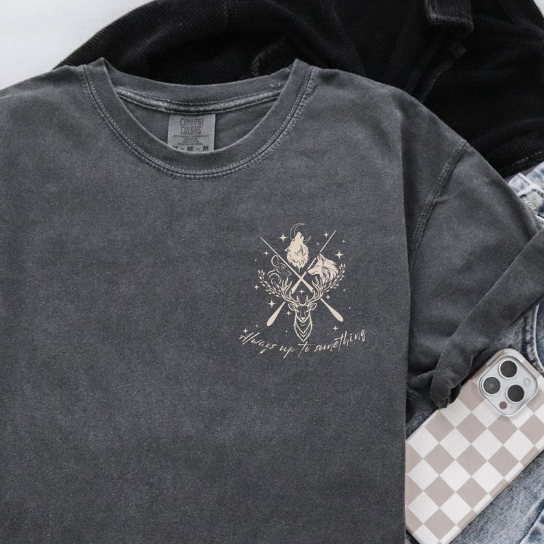 May include: Dark grey long-sleeve shirt featuring a cream-coloured graphic of a deer head, crossed oars, and the text "Always up to something". The shirt is made of a soft, comfortable material.