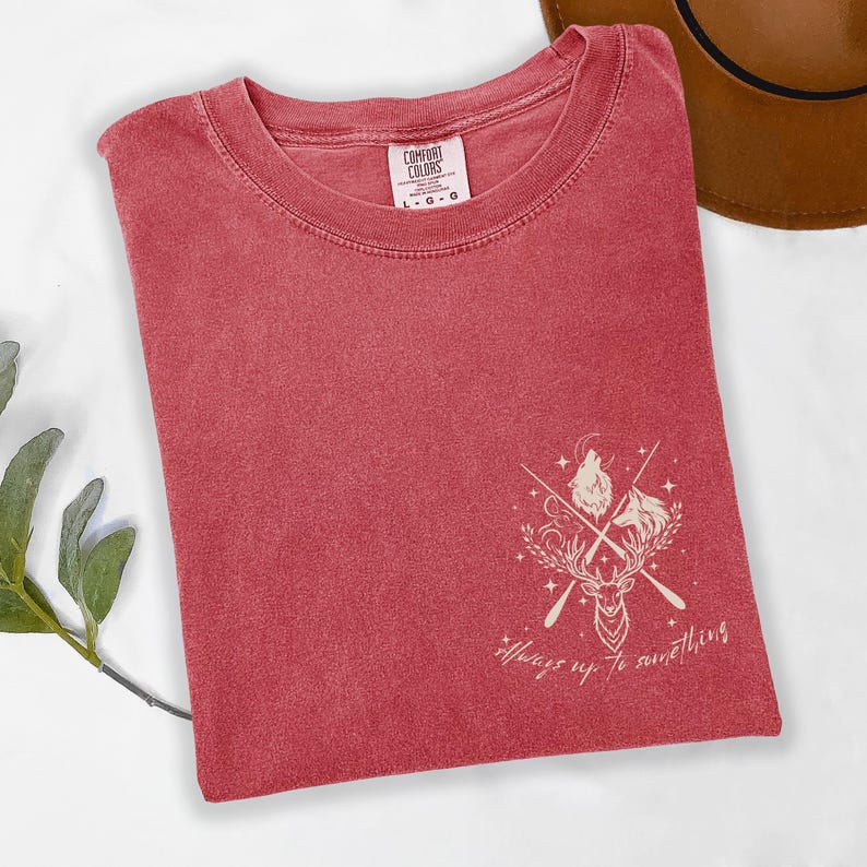 May include: A red Comfort Colors t-shirt featuring a deer, wolves, and crossed arrows graphic, with the text "Always up to something". The folded shirt has a tag at the top. The shirt is red.