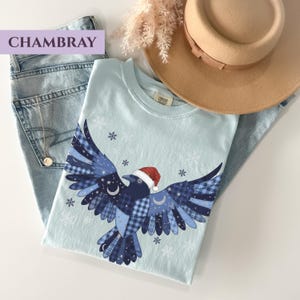 May include: A light blue t-shirt featuring a crow design with a Santa hat, snowflakes, and celestial accents. The crow's wings are patterned with blue plaid and stars. The shirt is paired with light wash jeans and a tan hat.