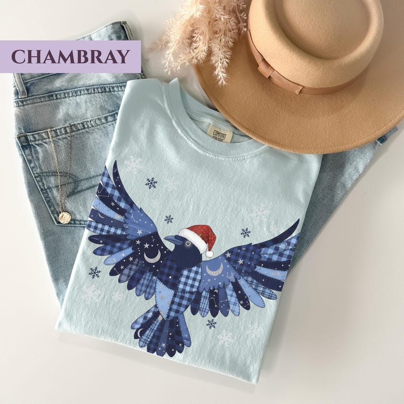 May include: A light blue t-shirt featuring a crow design with a Santa hat, snowflakes, and celestial accents. The crow's wings are patterned with blue plaid and stars. The shirt is paired with light wash jeans and a tan hat.