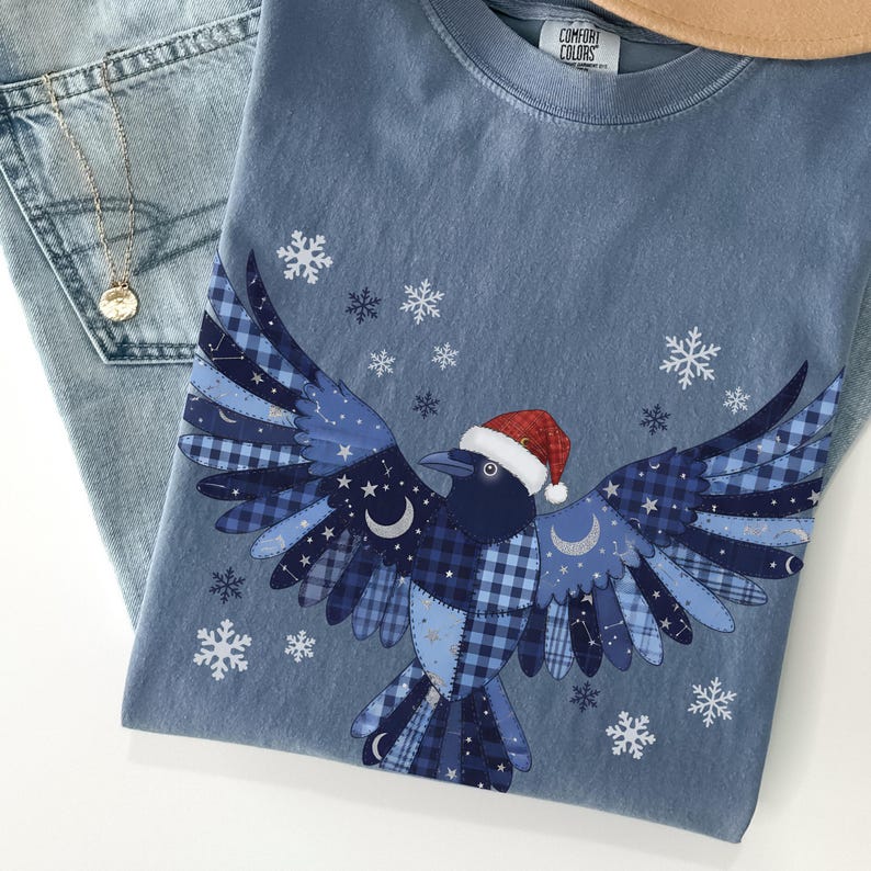 May include: A blue Comfort Colors t-shirt featuring a crow design with outstretched wings, wearing a Santa hat. The crow's wings are a patchwork of blue patterns, with snowflakes scattered around the design. A gold necklace and blue jeans are also visible.