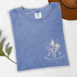 May include: A blue Comfort Colors t-shirt with a graphic design featuring a deer, wolf, and crossed arrows. The text "Always up to something" is below the graphic. A brown hat is visible in the background.