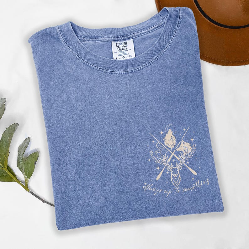 May include: A blue Comfort Colors t-shirt with a graphic design featuring a deer, wolf, and crossed arrows. The text "Always up to something" is below the graphic. A brown hat is visible in the background.