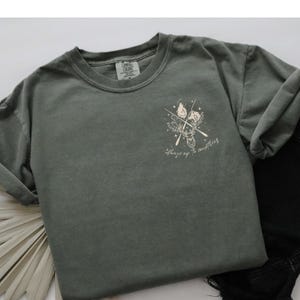 May include: A sage green t-shirt featuring a deer graphic, crossed arrows, and the text "Always up to something." The shirt is a Comfort Colors brand.