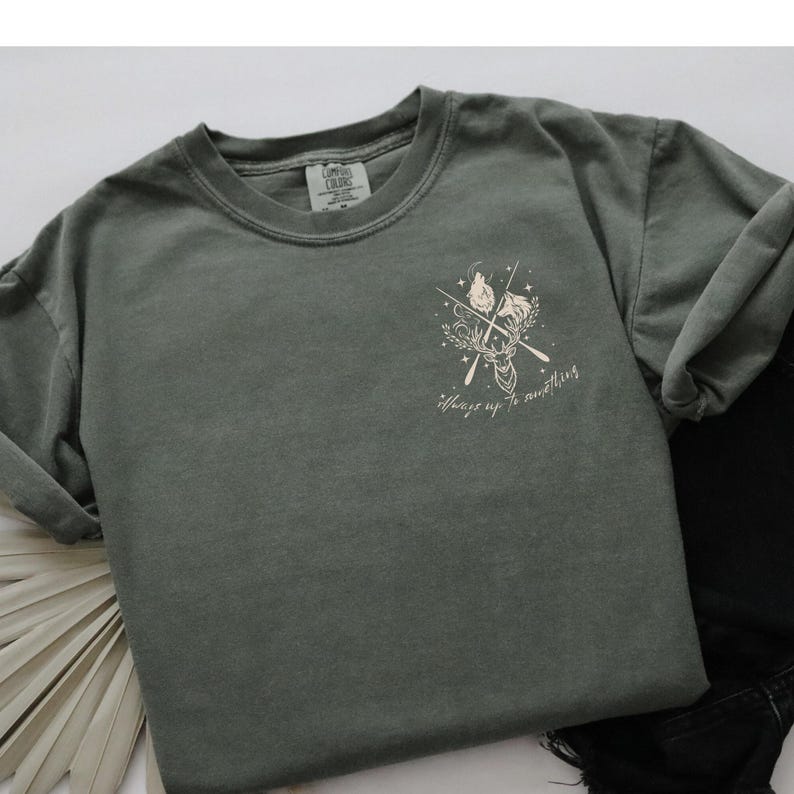 May include: A sage green t-shirt featuring a deer graphic, crossed arrows, and the text "Always up to something." The shirt is a Comfort Colors brand.