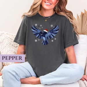 May include: Dark gray t-shirt featuring a blue and white patterned bird design with a Santa hat and snowflakes. The bird has outstretched wings and a crescent moon detail. The word "PEPPER" is in a light purple rectangle.