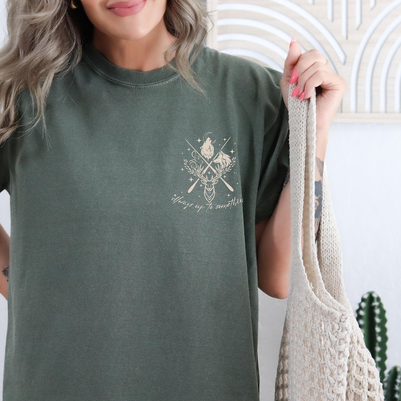 May include: Dark olive green t-shirt with a small chest graphic of a deer head, crossed arrows, and the text "Always up to something." The shirt has short sleeves and a crew neck.