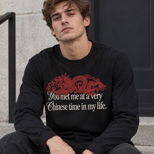 Puede incluir: Una camiseta negra de manga larga con un gráfico de dragón rojo sobre el texto blanco que dice: You met me at a very Chinese time in my life. La prenda tiene un cuello redondo y se combina con unos vaqueros de mezclilla gris oscuro.