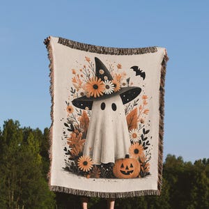 Puede incluir: Una manta de Halloween de color crema con un fantasma que lleva un sombrero de bruja adornado con flores. También se representa una calabaza y un murciélago. La manta tiene un borde con flecos y se sostiene contra un cielo azul y árboles verdes.