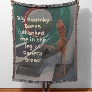 May include: A woven throw blanket featuring a cartoonish figure with the text "Big Badinky Bones shanked me in the leg at Panera Bread." The blanket has a fringed edge and a color palette of teal, gray, brown, and red.