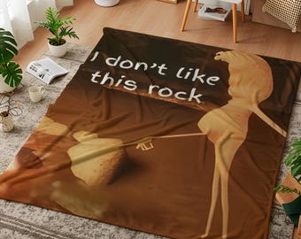 Può includere: Una coperta marrone con la scritta "I don't like this rock" in bianco. La coperta presenta un'illustrazione a cartoni animati di una persona che arrostisce un marshmallow sopra un fuoco. La coperta è realizzata in un materiale morbido.