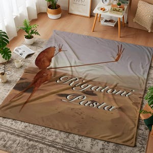 May include: A large, rectangular blanket with a desert-themed design. The blanket features a brown creature with outstretched arms and the words "Rigatoni Pasta" in a stylized font. The colour palette includes shades of brown and beige.