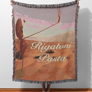 Può includere: Una coperta tessuta con un design di paesaggio desertico nei toni del marrone, beige e rosa. La coperta presenta le parole "Rigatoni Pasta" in un carattere decorativo. I bordi sono rifiniti con una frangia nera.