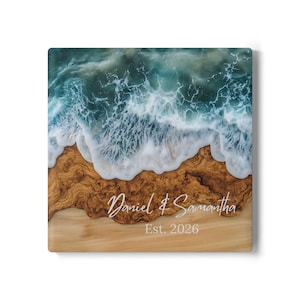 May include: Square wooden art piece depicting a beach scene. The top half shows a turquoise ocean with white foamy waves. The bottom half features a wooden beach with the names "Daniel & Samantha" and the year "Est. 2026" in white script.