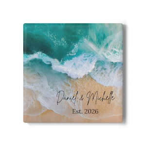 Può includere: Elemento decorativo quadrato con una scena di spiaggia con acqua turchese e onde bianche. I nomi "Daniel & Michelle" sono scritti in corsivo, con "Est. 2026" sotto. L'opera d'arte ha una finitura lucida.