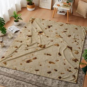 May include: A large, beige blanket with a pattern resembling a giant tortilla. The blanket has irregular brown spots and a soft, textured appearance. It is spread out on a patterned rug, with plants and furniture in the background.