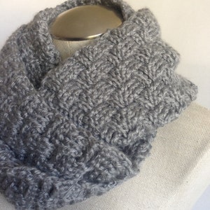 May include: A gray knitted infinity scarf with a textured pattern. The scarf is displayed on a white mannequin.