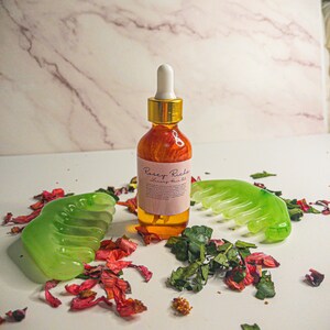 May include: A glass bottle of hair oil with a gold cap and a dropper. The bottle is labeled "Pursey Riches Luxury Hair Oil". The bottle is surrounded by green leaves and pink flower petals.