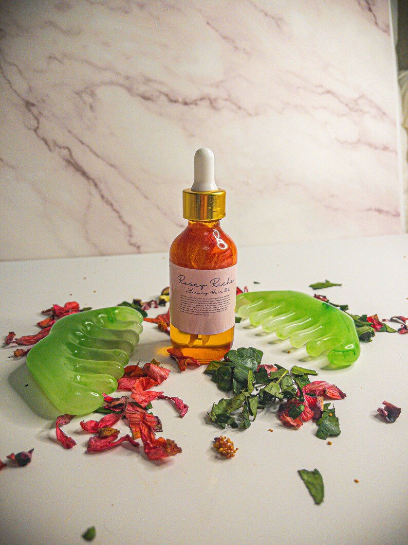 May include: A glass bottle of hair oil with a gold cap and a dropper. The bottle is labeled "Pursey Riches Luxury Hair Oil". The bottle is surrounded by green leaves and pink flower petals.