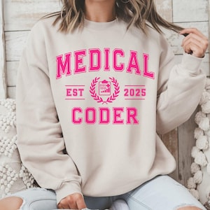 May include: A beige sweatshirt with the words "MEDICAL CODER" in bright pink, arched across the top and bottom. The text "EST 2025" is also in pink, with a medical symbol in the center. The sweatshirt is a casual style.