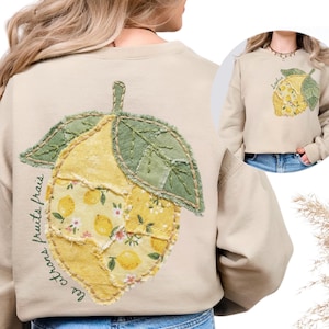 May include: Sand-colored sweatshirt featuring a large lemon applique on the back. The lemon design is a patchwork of yellow fabrics with floral and lemon patterns, and green leaves. The text "les citrons fruits frais" is printed on the left side of the lemon.