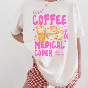 May include: Ivory-colored t-shirt with a pink and orange graphic that reads "iced COFFEE & MEDICAL CODER all day every day." The graphic includes an iced coffee, computer, stethoscope, and books.