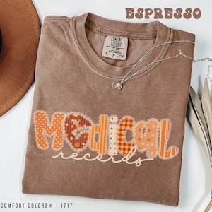 May include: Espresso-colored Comfort Colors t-shirt with the words "Medical Records" in a patchwork design. The letters are in shades of orange and brown with decorative stitching. A silver necklace with a pendant is draped on the shirt.
