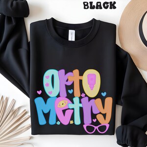 May include: Black crewneck sweatshirt with the word "Optometry" in colorful, bubble letters. The design includes hearts, flowers, and eyeglasses. The word "BLACK" is printed at the top of the image.