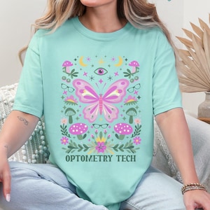 May include: A light teal Comfort Colors t-shirt featuring a colorful butterfly design with mushrooms, glasses, and the text "OPTOMETRY TECH." The design includes pink, purple, and green elements. The words "Chalky Mint" are written at the top.