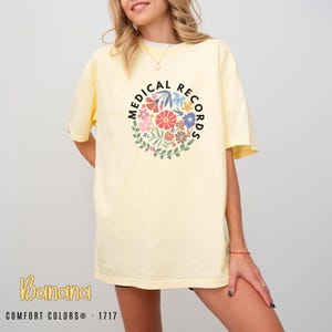May include: A light yellow t-shirt with the words "MEDICAL RECORDS" in a black circle around a floral design. The shirt also has the word "Banana" and "COMFORT COLORS® - 1717" printed on it.