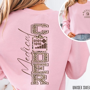 May include: A light pink unisex sweatshirt with the words "Medical Coder" in a vertical design on the back. The letters are brown with a leopard print pattern. The front of the sweatshirt has a smaller design with the words "Medical Coder".