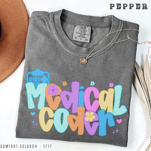 May include: A gray Comfort Colors t-shirt with the word "Medical Coder" in colorful, retro-style lettering. The shirt features a medical symbol graphic and small flower accents. A silver necklace with a pendant is draped on the shirt.