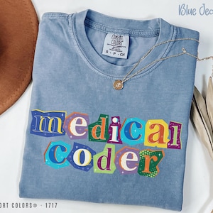 May include: A blue jean Comfort Colors t-shirt with the words "medical coder" in colorful, cut-out style letters. A gold necklace with a pendant rests on the shirt. The shirt is size S-CH.