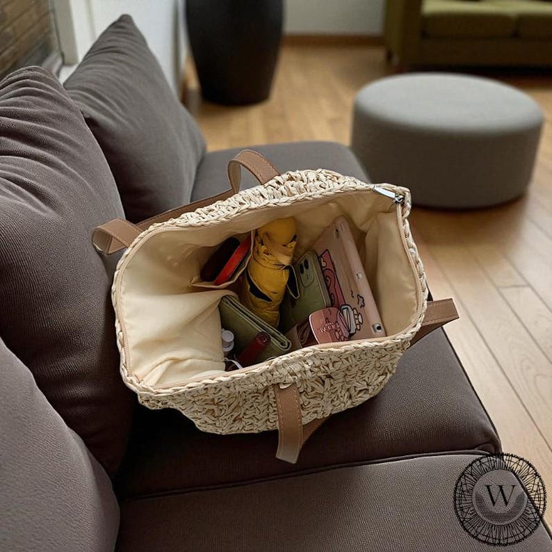 Puede incluir: Un bolso tote de paja tejida con asas de cuero marr&oacute;n y un interior beige. El bolso est&aacute; abierto, revelando un paraguas amarillo, carteras y tel&eacute;fonos inteligentes. El bolso est&aacute; sobre un sof&aacute; marr&oacute;n.