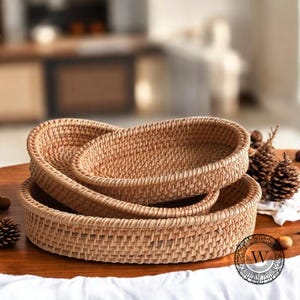 Puede incluir: Tres cestas ovaladas tejidas de diferentes tamaños, hechas de ratán natural, apiladas sobre una superficie de madera. Las cestas son de color marrón claro y tienen un aspecto natural y rústico.