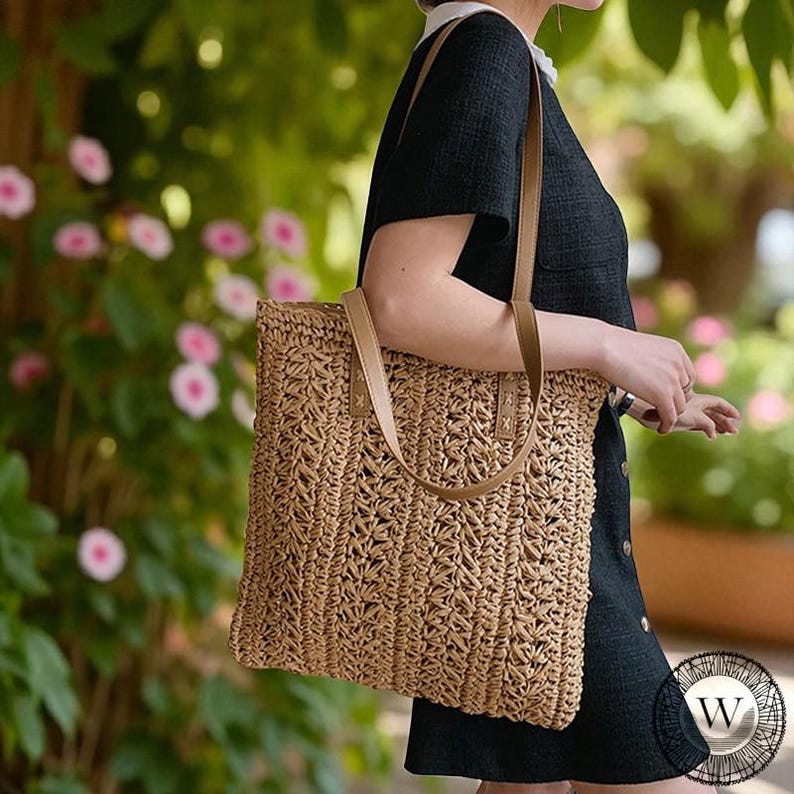 Puede incluir: Un bolso tote de paja tejida con asas de cuero marr&oacute;n claro. El bolso tiene forma rectangular y un patr&oacute;n texturizado. El bolso se lleva al hombro. El fondo presenta flores rosas y follaje verde.