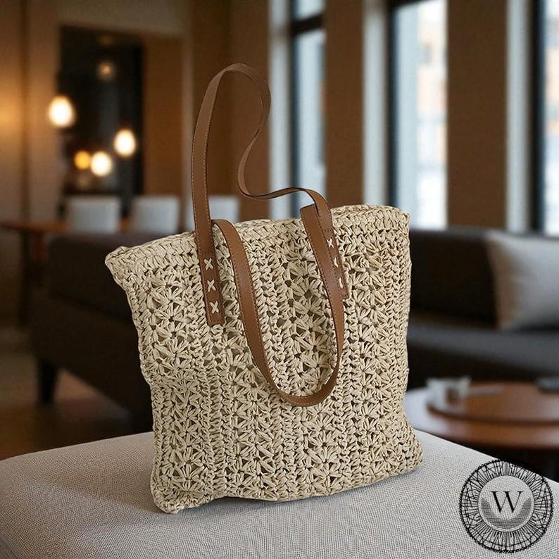 Puede incluir: Un bolso tote de paja tejida con asas de cuero marr&oacute;n. El bolso tiene forma rectangular y un dise&ntilde;o de tejido abierto y texturizado. Las asas est&aacute;n unidas con costuras decorativas. El bolso est&aacute; sobre una superficie gris claro.