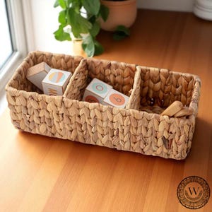 Puede incluir: Una cesta rectangular tejida con tres compartimentos, llena de bloques de madera y pequeños utensilios de madera. Los bloques tienen diseños geométricos coloridos y números. La cesta está hecha de fibras naturales y se encuentra sobre una superficie de madera.