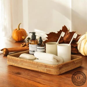 May include: A woven wicker tray with a bar of soap, two white towels, two glass bottles, two toothbrushes, and two ceramic cups. The tray is on a wooden surface with a autumnal background.