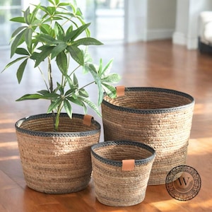 Puede incluir: Tres cestas tejidas de diferentes tamaños, hechas de cuerda de yute natural con un borde negro y asas de cuero. Las cestas están anidadas una dentro de la otra, con la cesta más grande en la parte inferior y la cesta más pequeña en la parte superior.