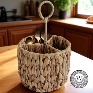 Può includere: Porta utensili in vimini con manico. Il cestino è diviso in tre sezioni e contiene diversi cucchiai color oro. Il cestino è realizzato con fibre naturali e poggia su una superficie in legno. Un emblema decorativo è sul lato destro.