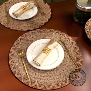 May include: A dining table setting featuring two woven placemats. Each place setting includes a white plate, a rolled beige napkin held by a gold ring, and gold cutlery. The placemats have a geometric border.