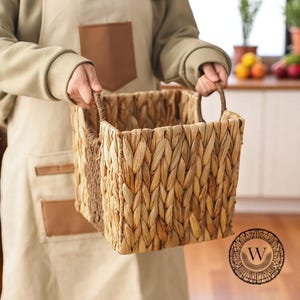 Può includere: Un cesto rettangolare intrecciato con manici viene tenuto in mano. Il cesto è realizzato in materiale intrecciato di colore naturale. Il cesto ha una forma quadrata ed è progettato per la conservazione. Lo sfondo include un ambiente cucina.