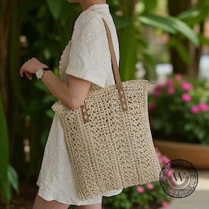 Puede incluir: Un bolso tote tejido beige con correas de cuero marr&oacute;n. El bolso tiene forma rectangular y cierre de cremallera. Las correas est&aacute;n unidas con costuras decorativas. El bolso se lleva al hombro.