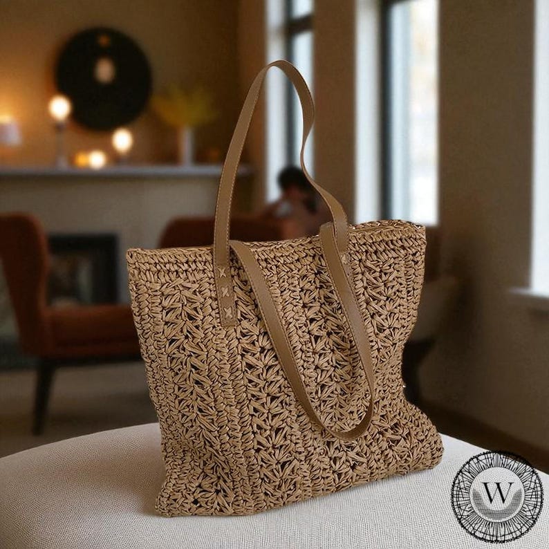Puede incluir: Un bolso tote de paja tejida con asas de cuero marr&oacute;n claro. El bolso tiene forma rectangular y un dise&ntilde;o de tejido abierto texturizado. Las asas est&aacute;n unidas con costuras decorativas. El bolso est&aacute; sobre una superficie blanca.